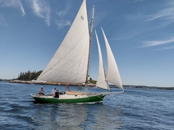 A sailboat with white sails is on a calm body of water with people on board, under a blue sky with wispy clouds in the distance by a small island.