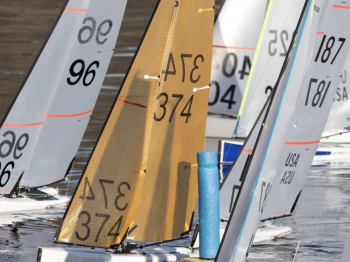 Several small, numbered sailboats float close together in the water, likely in a competitive event.
