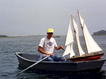 A person is sitting in a small wooden boat on the water, holding a model sailboat which is also in the boat.