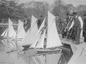 A group of people in vintage clothing gather around the edge of a pond to sail model boats.