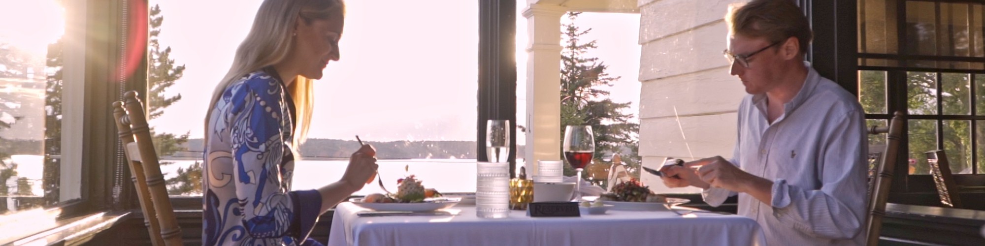 A couple is dining together at a table near large windows with a scenic view of water and trees during sunset.