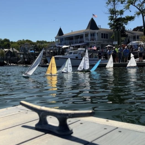 Small model sailboats are floating on the water near a dock, with a building and spectators in the background, under a clear sky.