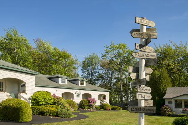 A landscaped yard with houses and a signpost indicating distances to various destinations, surrounded by trees under a clear blue sky.