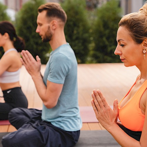 Three people are practicing yoga outdoors, sitting in a meditative pose with their hands in a prayer position, focusing on mindfulness and relaxation.