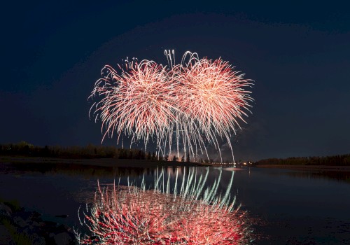 The image shows a nighttime scene of colorful fireworks exploding over a placid body of water, with reflections visible on the water's surface.