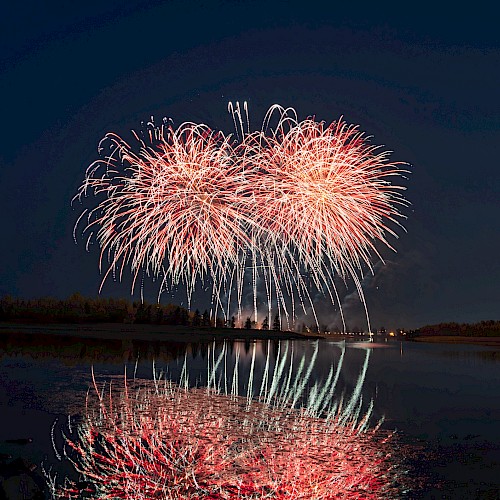 The image shows a nighttime scene of colorful fireworks exploding over a placid body of water, with reflections visible on the water's surface.