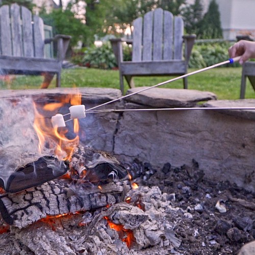 Two marshmallows being roasted over a campfire with chairs and greenery in the background.