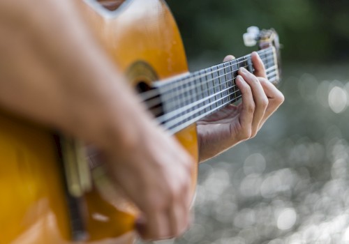 A person is playing an acoustic guitar, with focus on their hands and the guitar. The background features a blurred, natural outdoor setting.