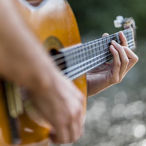 A person is playing an acoustic guitar, with focus on their hands and the guitar. The background features a blurred, natural outdoor setting.