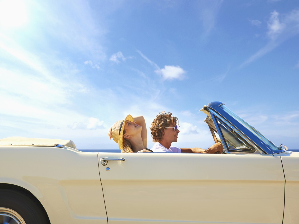 Two people are enjoying a sunny day in a convertible car, with the top down and blue skies overhead.