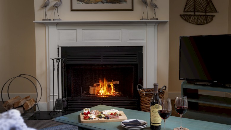 A cozy living room with a lit fireplace, a coffee table with red wine and snacks, and a TV on a stand.