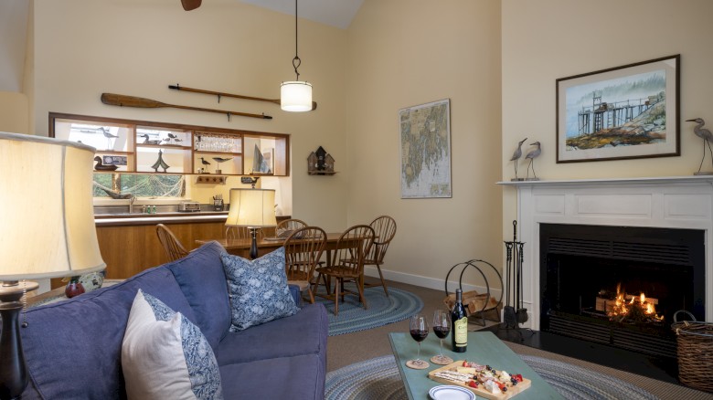 A cozy living room with a blue sofa, a fireplace, a coffee table with food and wine, and a dining area near the kitchen under a high ceiling.