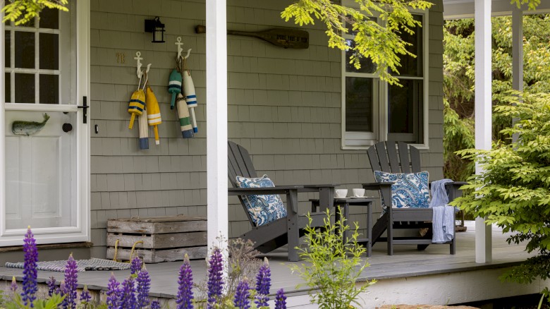 A cozy porch with two chairs, a small table, colorful hanging jackets, and a white door. Various plants and purple flowers are in front.