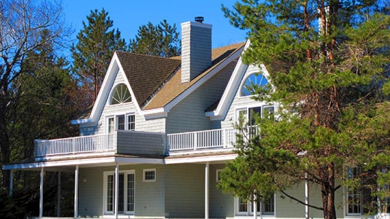 The image shows a gray two-story house with a unique roof design, two chimneys, a large balcony, and surrounded by trees and a clear blue sky.