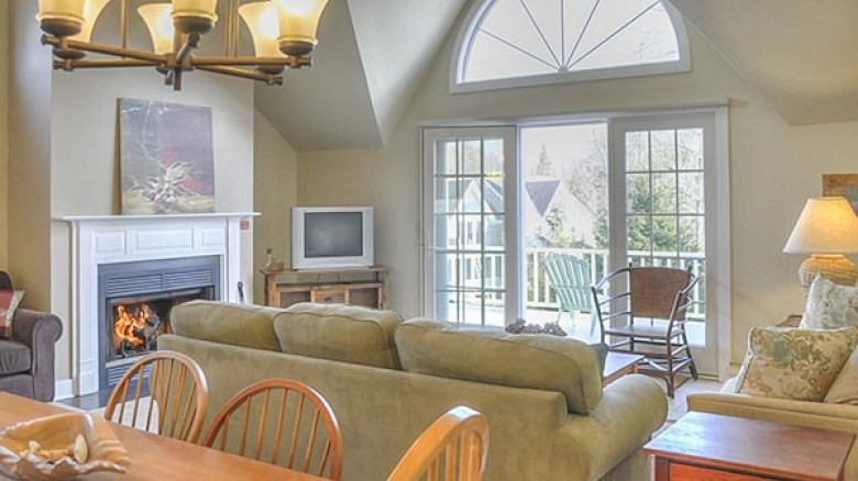 A cozy living room with a fireplace, TV, large windows, and a chandelier above the dining table. There is seating for multiple people.