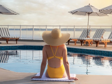 A person in a yellow swimsuit and wide-brimmed hat sits by a pool near lounge chairs and umbrellas, overlooking the water.