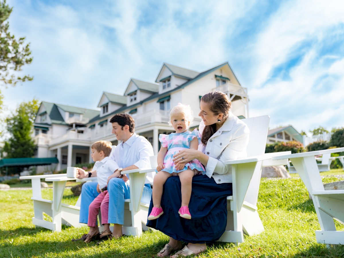 A family sits on chairs in a grassy area outside a large house on a sunny day.