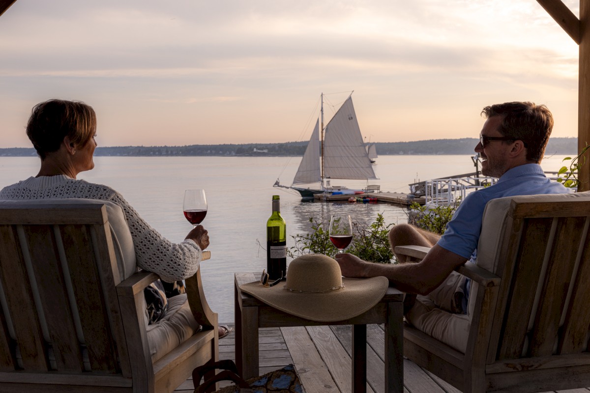 Two people relax with wine on a deck overlooking a serene lake, watching a sailboat glide by under a sunset sky.